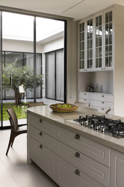 Detalhe de cozinha com ilha central em pedra clara, cooktop embutido e gavetas com puxadores tipo concha. Ao fundo, cristaleira com portas de vidro e armários inferiores. Janela ampla revela jardim interno com árvore e iluminação natural abundante.