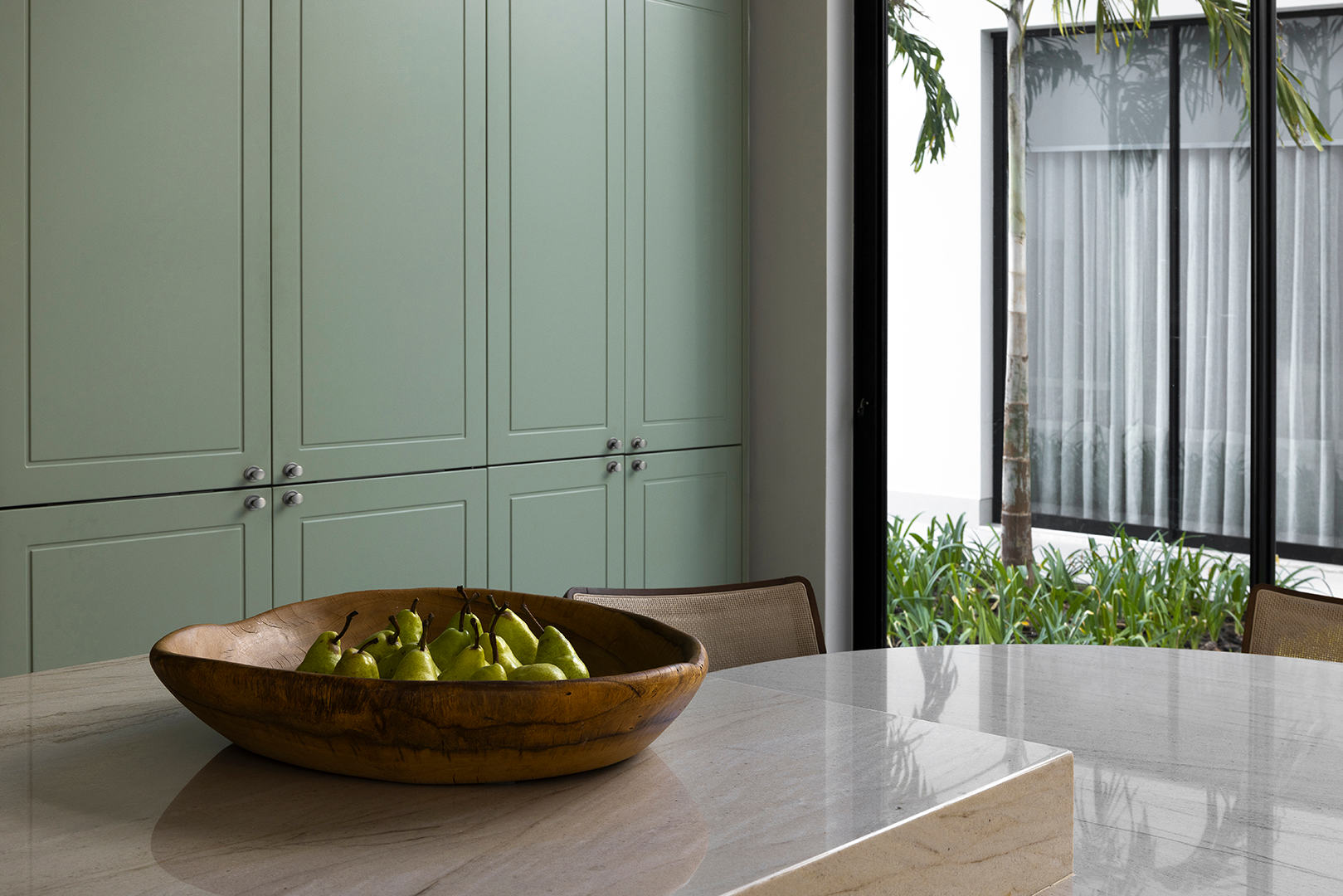 Armário verde com acabamento clássico em microtextura, com puxadores cromados duplos, ao fundo de uma cozinha elegante com bancada em quartzito arena e fruteira de madeira com peras verdes. Ao lado, grande janela com vista para jardim externo.