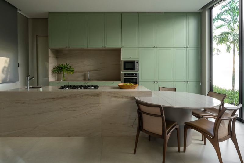 Cozinha com marcenaria verde em acabamento clássico e microtextura, equipada com forno embutido, cooktop e armários com puxadores cromados. Ilha central e bancada em quartzito com mesa acoplada, cadeiras de madeira e palhinha, ao lado de ampla janela com vista para jardim externo."