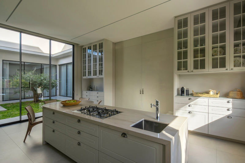 Cozinha com ilha central em pedra clara, armários em tom neutro com puxadores tipo concha e cristaleiras com portas de vidro. Ao fundo, ampla porta de vidro com vista para jardim interno com árvore central, trazendo iluminação natural para o ambiente.